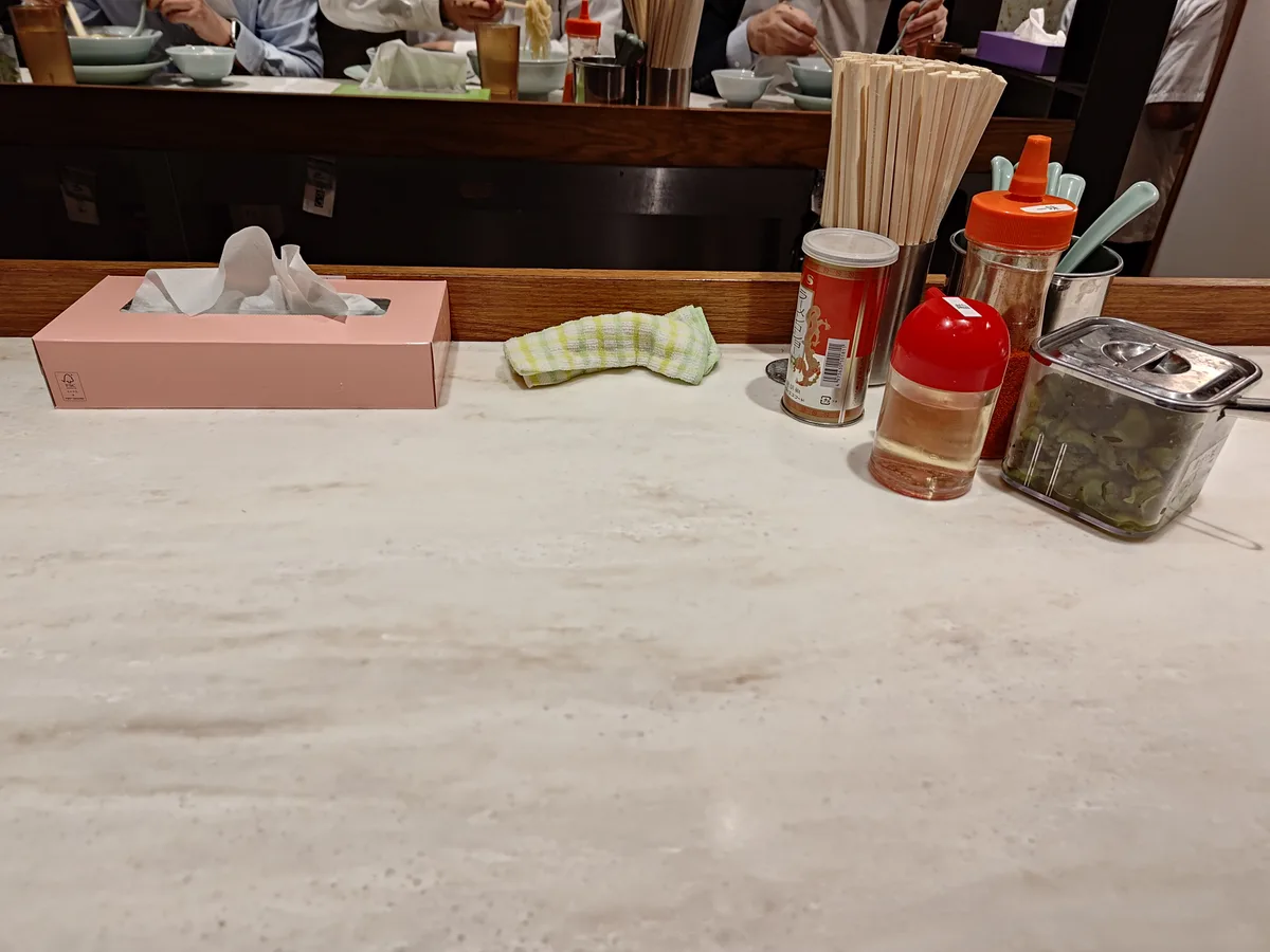 Würzmittel auf dem Tisch bei Chiechan Ramen Kanda: Pfeffer, Essig, Chili, frischer Knoblauch und eingelegtes Gemüse für den kostenlosen Reis. Perfekt für Geschmacksveränderungen der Chan-kei Ramen.