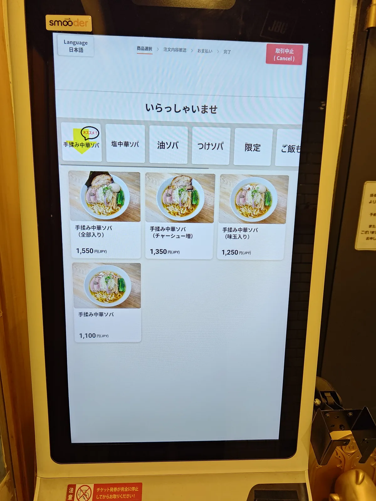 Ticketautomat des Chūka Soba Billiken. Ein smooder-Touchpanel-Modell mit einer 'Language 日本語'-Sprachwechselschaltfläche oben links, einer Fortschrittsanzeige '商品選択 → 注文内容確認 → お支払い → 完了' oben, und Menükategorien-Tabs an der linken Seite (手揉み中華ソバ・塩中華ソバ・油ソバ・つけソバ・限定・ご飯も…). In der Mitte werden die vier Hauptgerichte des 'Temomi Chūka Soba' (Zenbu-iri ¥1.550 / Chashu-ma. ¥1.350 / Ajitama-iri ¥1.250 / Plain ¥1.100) mit Fotos angezeigt. A Japanese ramen ticket machine with multilingual support for foreign tourists.