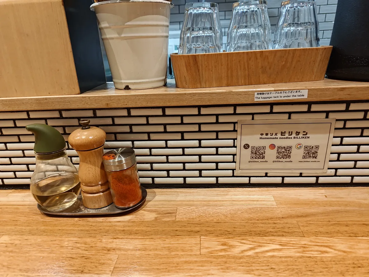 Inneneinrichtung des Billiken Ramen-Shops: Eine saubere Holztheke mit Gewürzen, Wasser und Gläsern zur Selbstbedienung. Westernmusik spielt im Hintergrund, und es sind nur wenige Gäste da, was viele freie Plätze lässt. A modern and clean Japanese ramen counter interior, perfect for enjoying a meal.