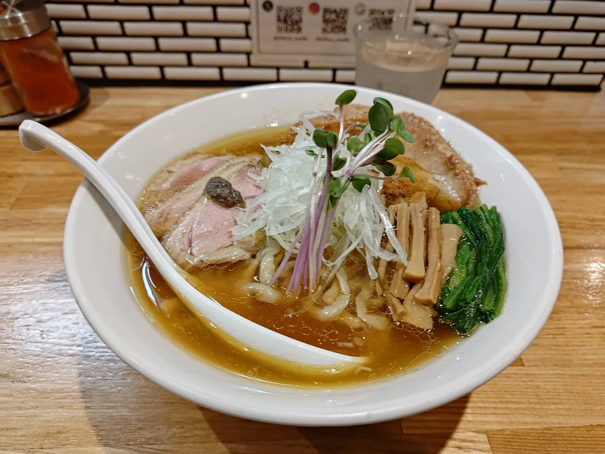 Das Temomi Chūka Soba (Chashu-ma.) von Billiken, serviert in einer weißen Schale. Eine klare, bernsteinfarbene Shoyu-Suppe, in der handgeknetete, gewellte Nudeln schwimmen. Toppings sind bei niedriger Temperatur gegartes Schweine-Chashu, gerösteter Entenbraten mit schwarzer Trüffelpaste, Spinat, Menma und weißer Lauch, appetitlich angerichtet. Japanese ramen bowl with duck and pork chashu in a clear broth.