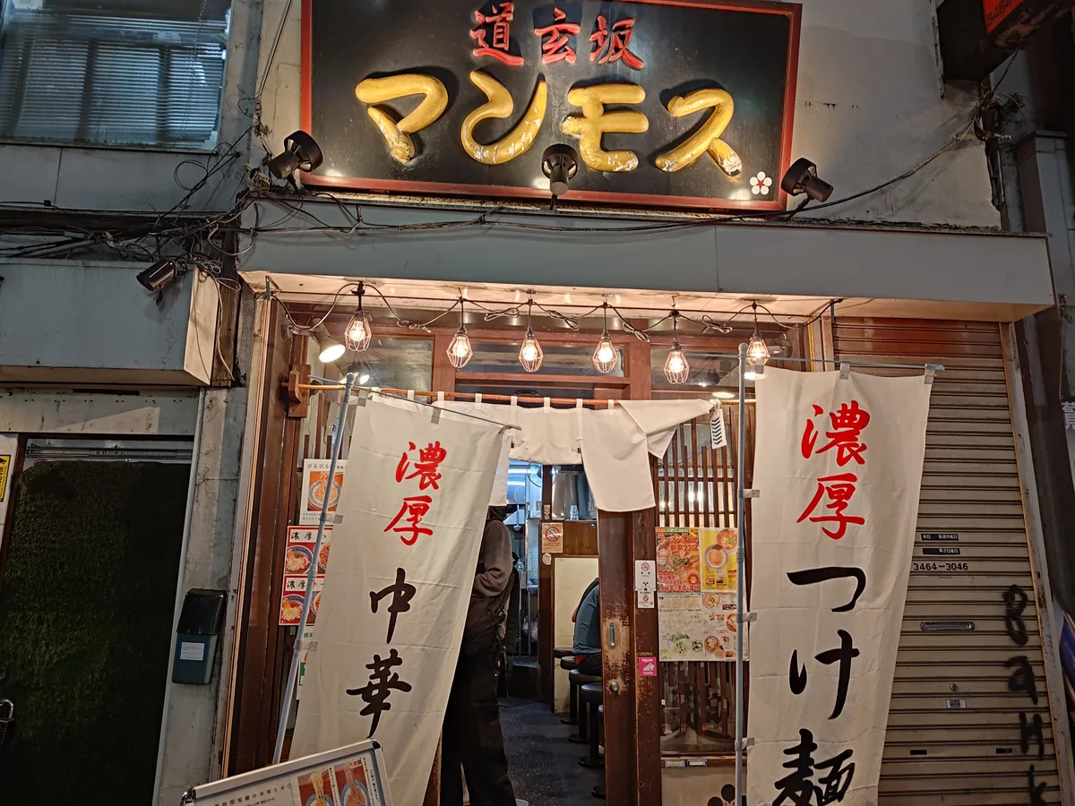 Dogenzaka Manmosu ramen shop exterior in Shibuya, Japan. A golden sign and glowing noren mark the entrance. White banners display 'Rich Chuka' and 'Rich Tsukemen,' highlighting the specialties of this popular Japanese ramen spot.