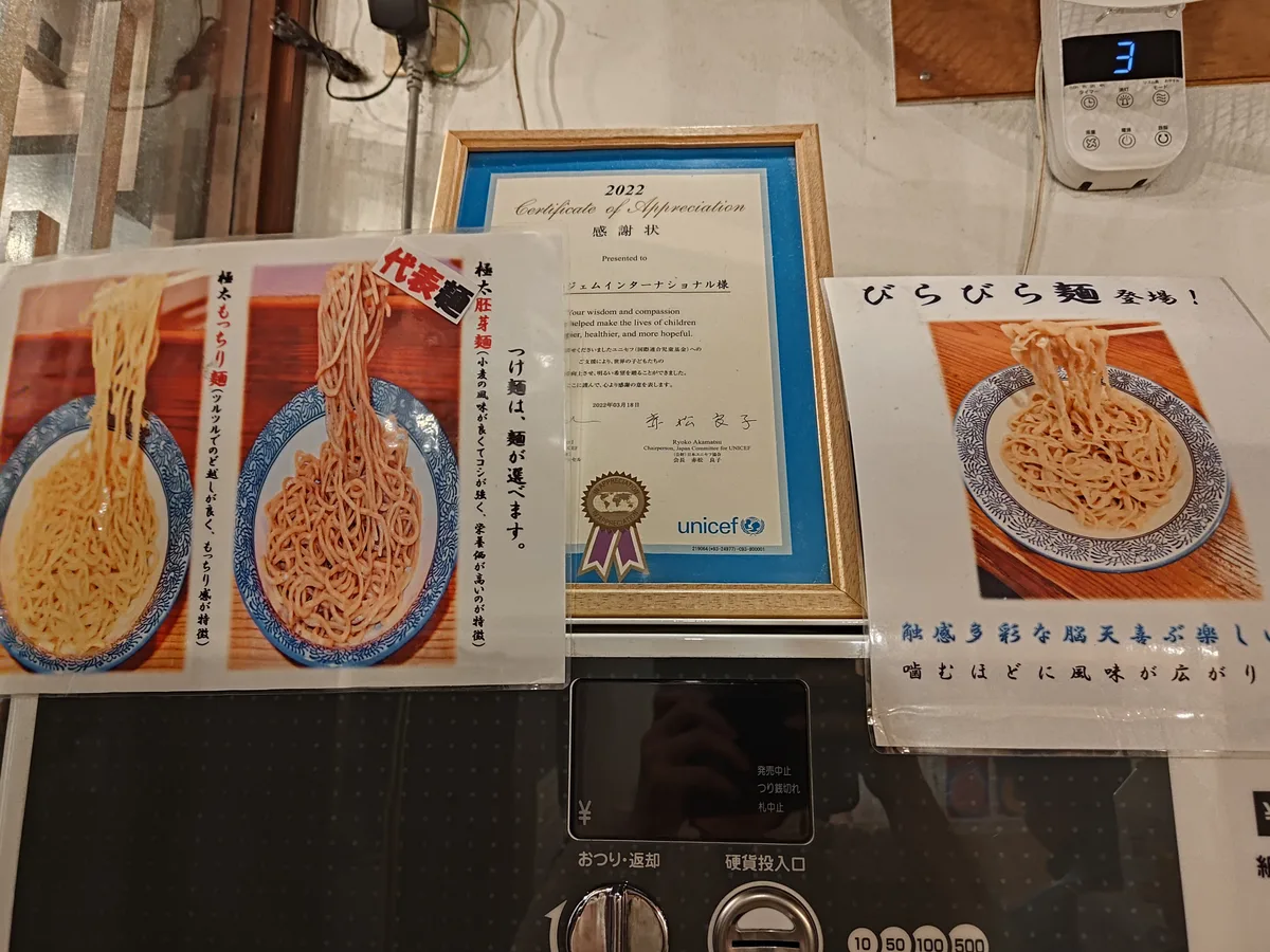 Noodle options at Dogenzaka Manmosu, a ramen shop in Shibuya, Japan. A poster shows 'Futomen Mocchiri-men,' 'Futomen Haigamen,' and 'Birabira-men' to help customers choose their preferred ramen noodle type.