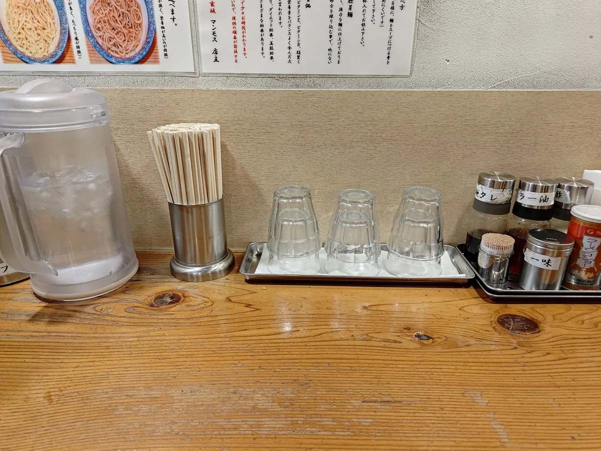 Clean counter at Dogenzaka Manmosu, a Japanese ramen shop in Shibuya. Features a cold water pot, chopsticks, glasses, and condiments like kashi, chili oil, and ichimi. This setup is typical for a ramen shop in Japan.