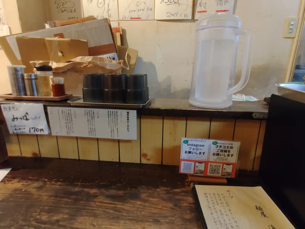 店內吧台的景象。上方擺放著水壺、水杯和調味料，燈光沉靜的空間。日本新宿、拉麵店、內部裝潢、舒適