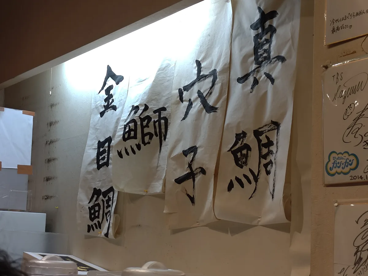 店內牆上以毛筆字張貼的「本日的魚身精華」。金目鯛、鰤魚、星鰻、真鯛四種魚名清晰可見。日本新宿拉麵、麵屋海神、每日限定、魚骨高湯