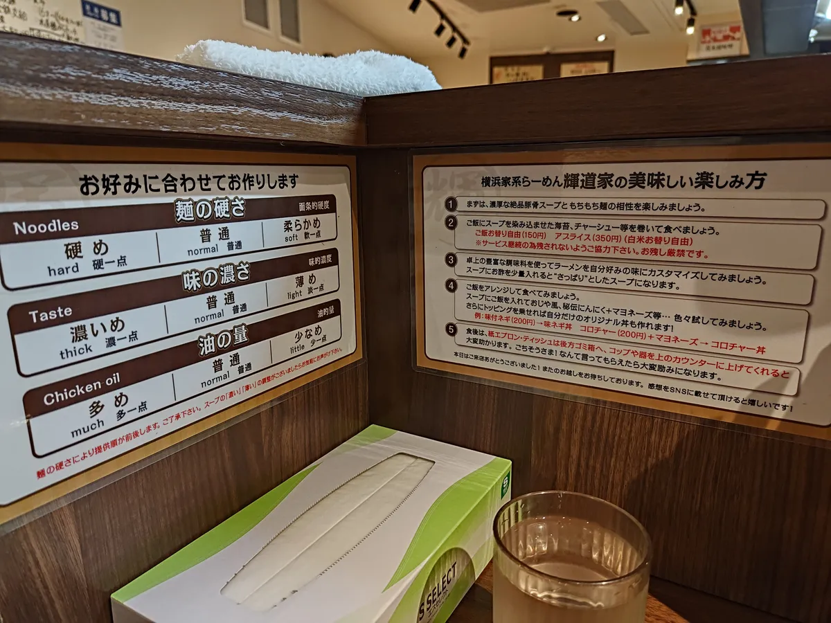 カウンターに設置されたお好みカスタマイズ表。麺の硬さ（Noodles）、味の濃さ（Taste）、油の量（Chicken oil）が日英併記で案内されている。右側には横浜家系らーめん輝道家の美味しい楽しみ方が書かれている。