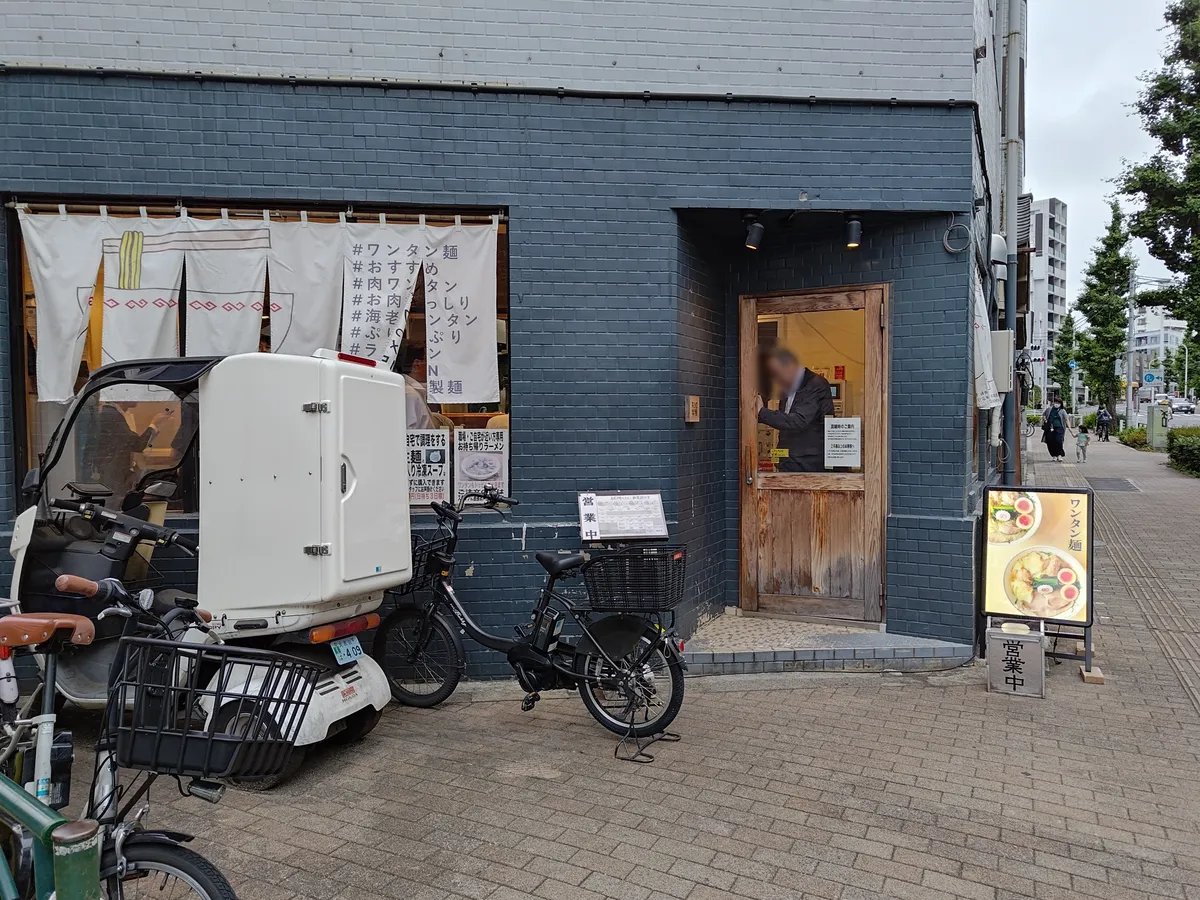 キング製麺の店舗外観。濃紺のレンガ調の壁に木製の引き戸、左手の窓にはラーメン丼を描いた白い暖簾と「ワンタン麺」などの幟、右手に「営業中」のA型看板が立つ。
