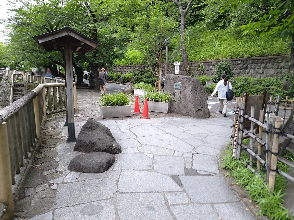 北区立音無親水公園の石畳の遊歩道。両側に石垣と竹垣、奥には新緑の木々が茂り、散策する人の姿が見える。