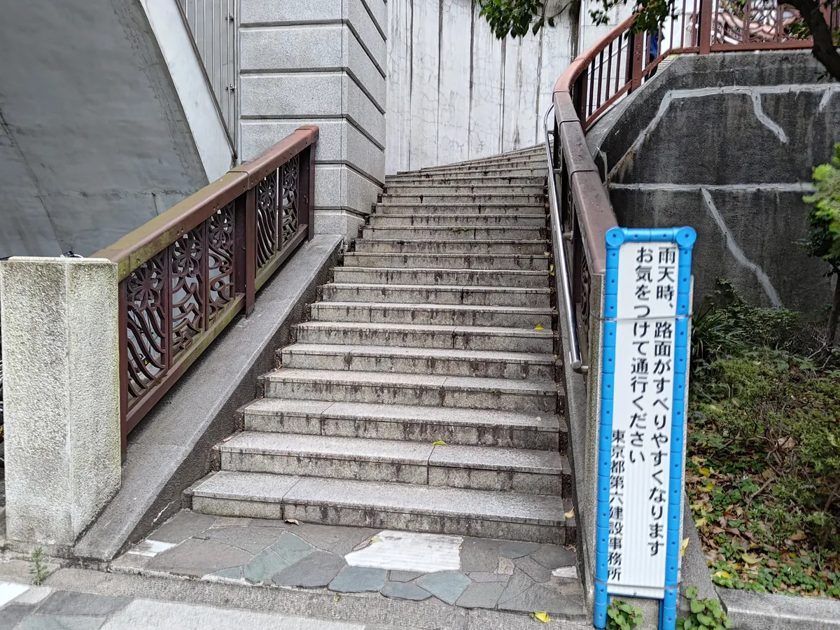 陸橋へ上がるコンクリートの階段。装飾のある手すりが付き、脇に「雨天時、路面がすべりやすくなります お気をつけて通行ください」と書かれた青い看板が立つ。