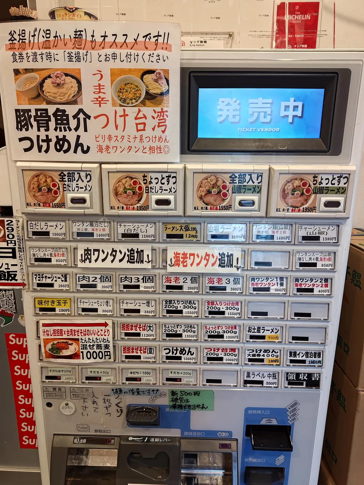 キング製麺のタッチパネル式券売機。画面に「発売中（TICKET VENDOR）」と表示され、右側に料理写真付きのボタンが多数並ぶ。左には豚骨魚介つけめんなどのポスターが貼られている。