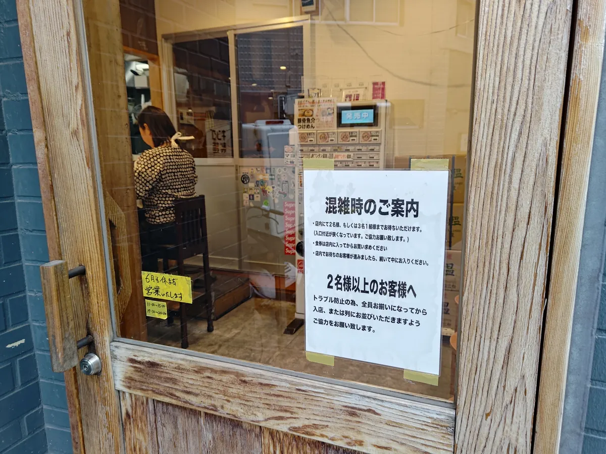 入口ドアに貼られた「混雑時のご案内」の張り紙。前の客が着席してから食券を購入すること、2名以上は全員そろってから入店・整列することなどが日本語で書かれている。ガラス越しに店内の券売機とカウンター席が見える。