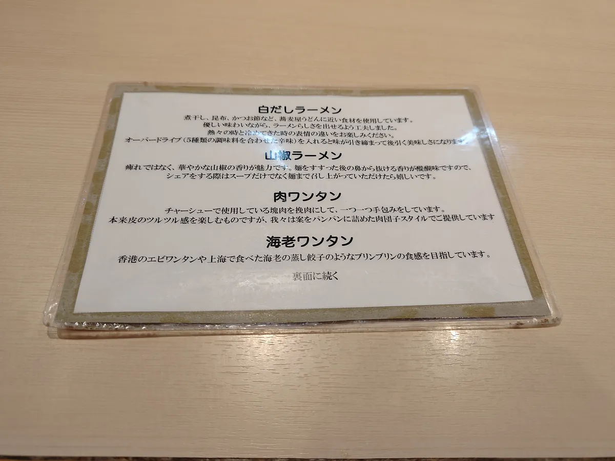 カウンターに置かれたラミネート加工のメニュー説明カード。白だしラーメン、山椒ラーメン、肉ワンタン、海老ワンタンそれぞれの特徴が日本語で説明されている。
