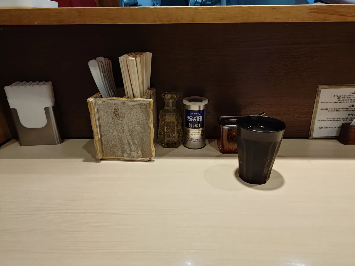 キング製麺のカウンター卓上。紙ナプキン、箸とレンゲの入れ物、酢の瓶、S&Bの白胡椒、唐辛子の小瓶、黒いタンブラー（水用）、右手にメニュー説明カードが並ぶ。
