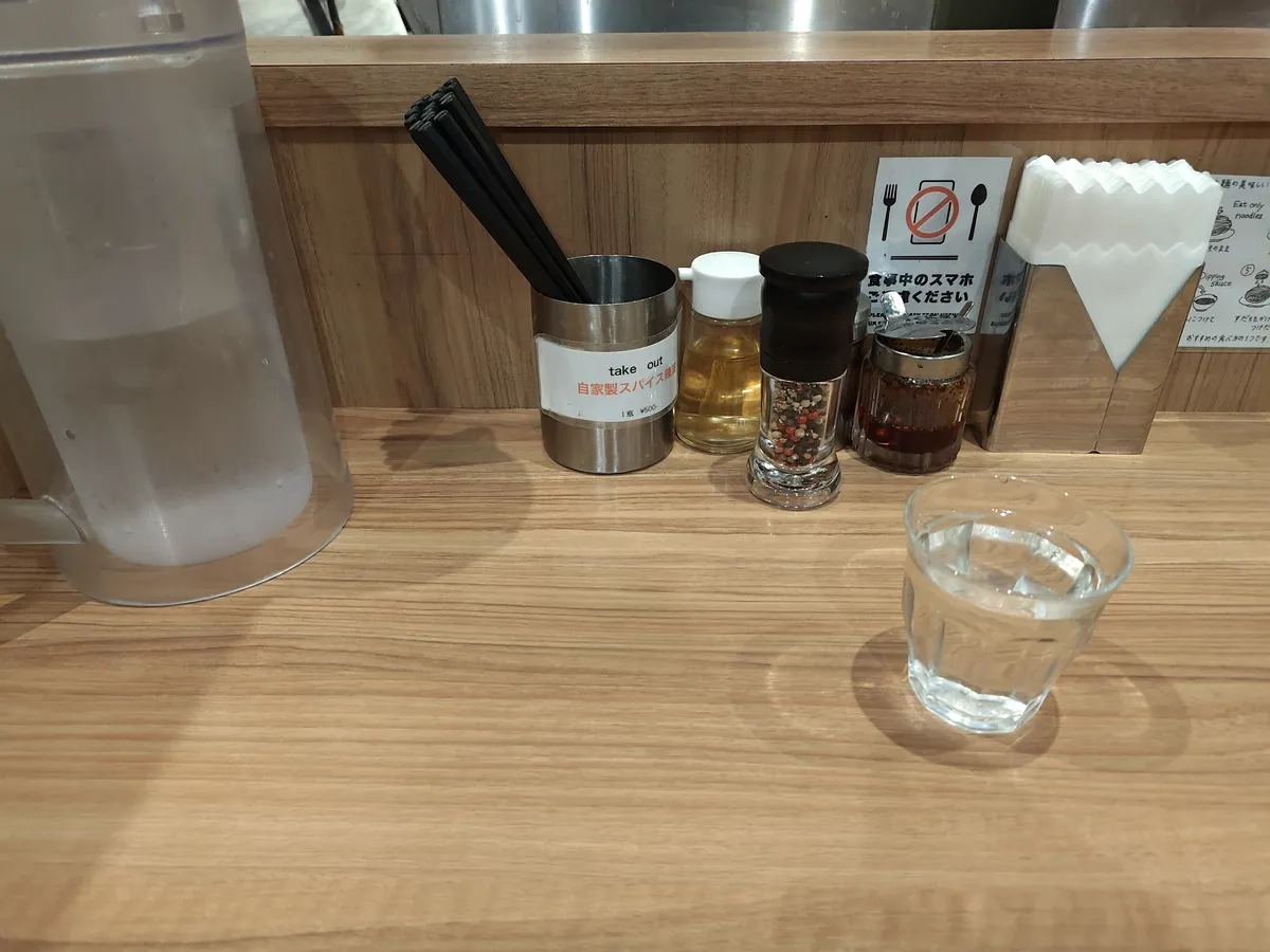 カウンター席の卓上。水のグラスとピッチャー、自家製スパイス、ペッパーミル、酢、唐辛子などが並ぶ。奥に「食事中のスマホはご遠慮ください」の張り紙が見える。