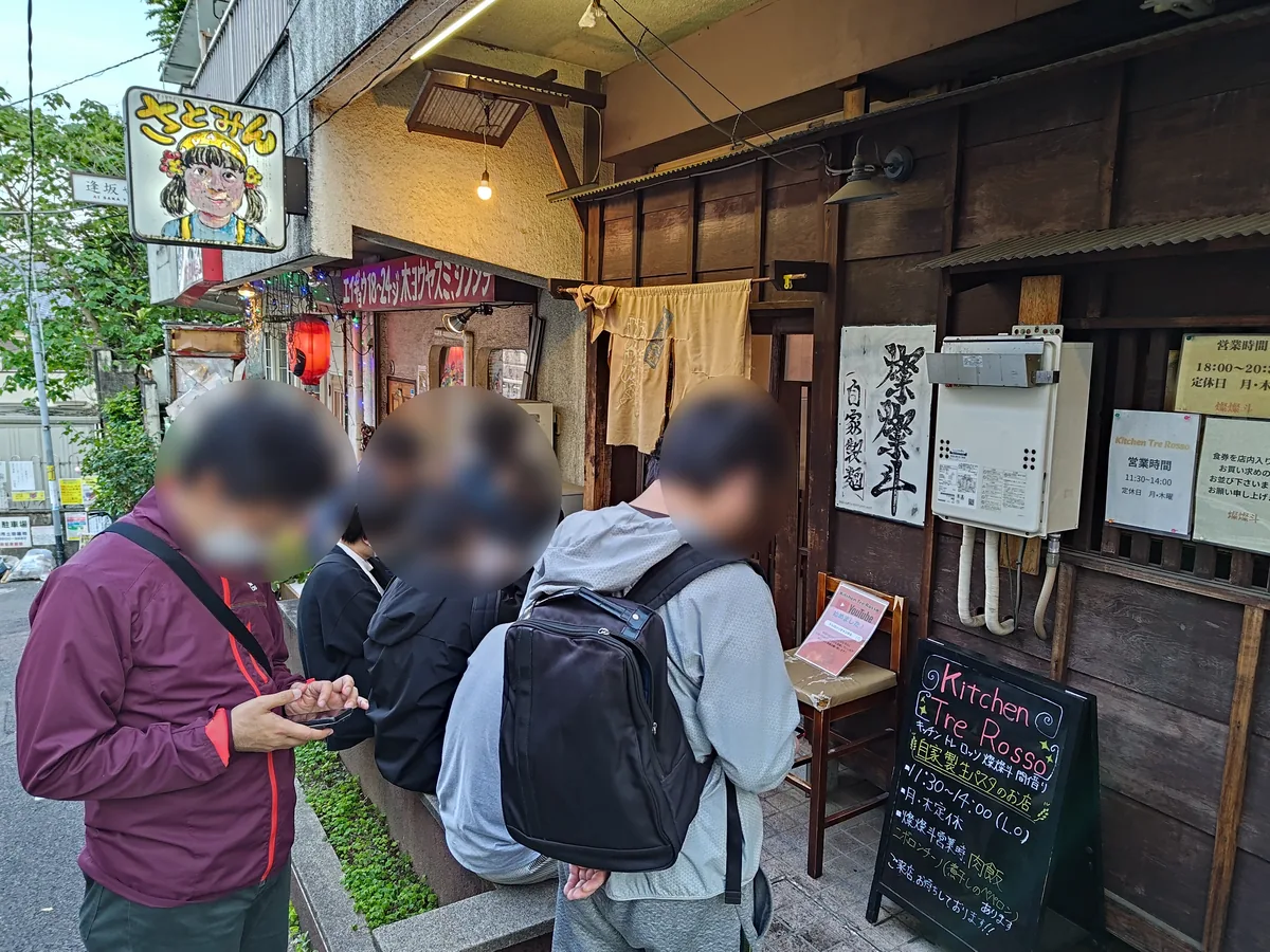 燦燦斗の店舗外観。木目調の店構えに使い古された黄色い暖簾と「燦燦斗」の墨書き、入り口前には数名の客が並ぶ。隣には昼に親族が営むイタリアン「Kitchen Tre Rosso」の黒板看板も見える。
