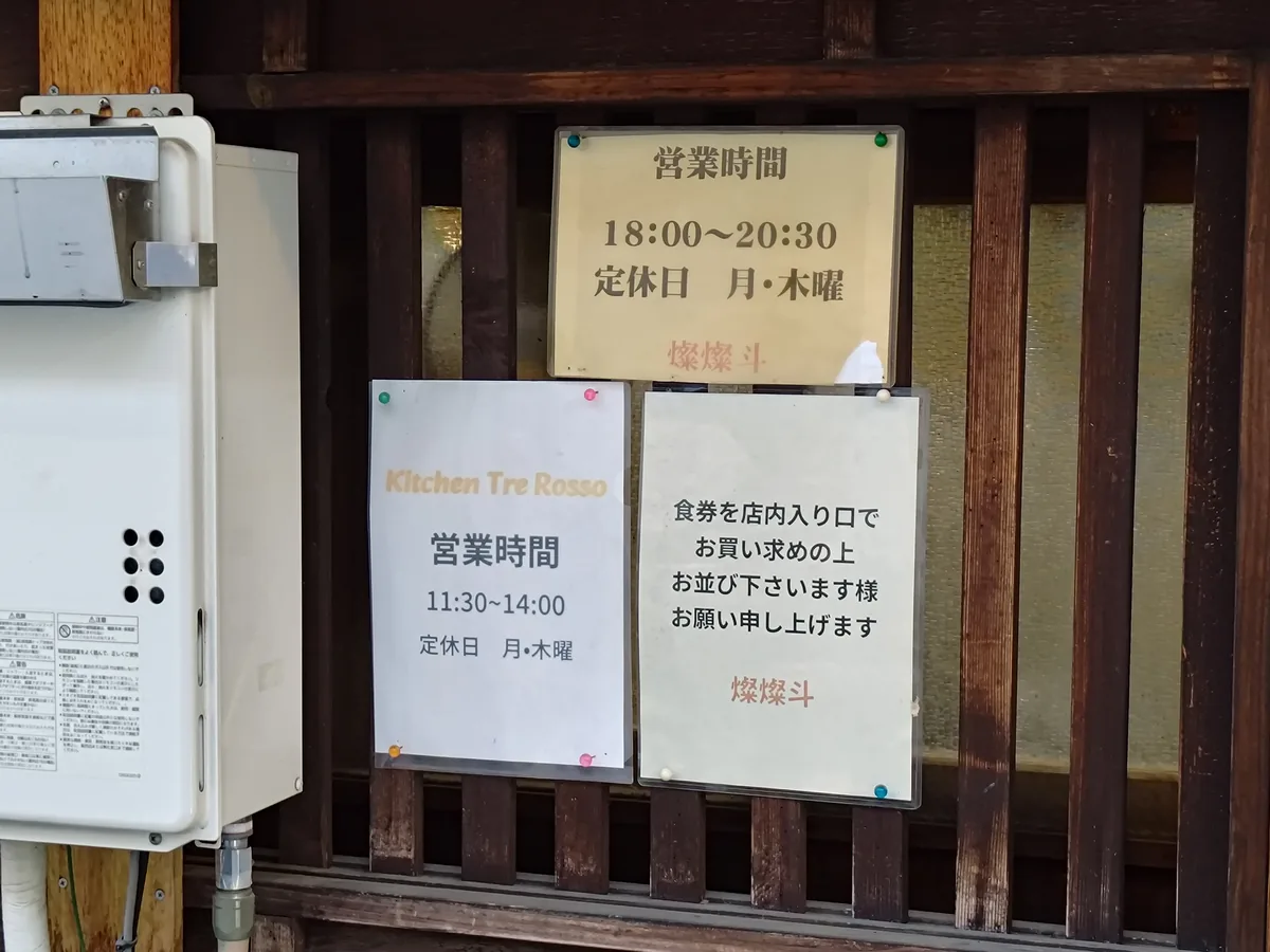 店頭に掲示された営業時間の張り紙。「営業時間 18:00〜20:30 定休日 月・木曜」と「燦燦斗」の朱印。同じ場所で昼に営業する「Kitchen Tre Rosso」（11:30〜14:00、月木休）の張り紙も並ぶ。