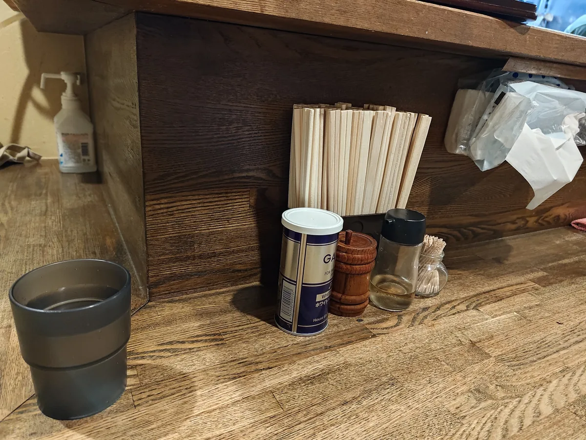 カウンター上段に並ぶ卓上調味料。割り箸入れ、白胡椒（GABAN）、木製の七味入れ、ガラスの調味料瓶、消毒スプレー、グラスなどがコンパクトに配置されている。