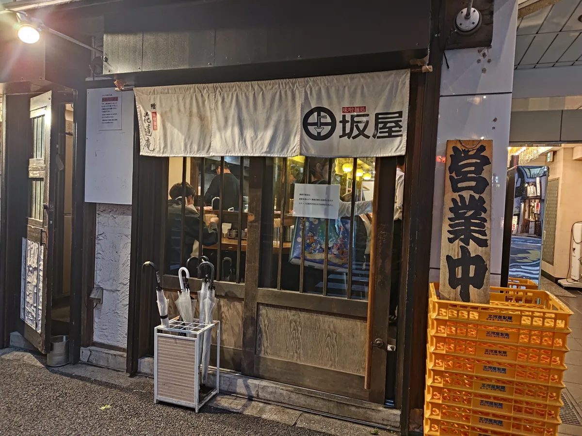 Miso Mendokoro Tasakaya 的夜間外觀。白色的暖簾上印有圓形標誌和「坂屋」字樣，右側有藍色的「營業中」招牌。木紋設計的店面沉穩雅致，店門口擺放著傘架。 東京池袋拉麵 獨特味噌口味