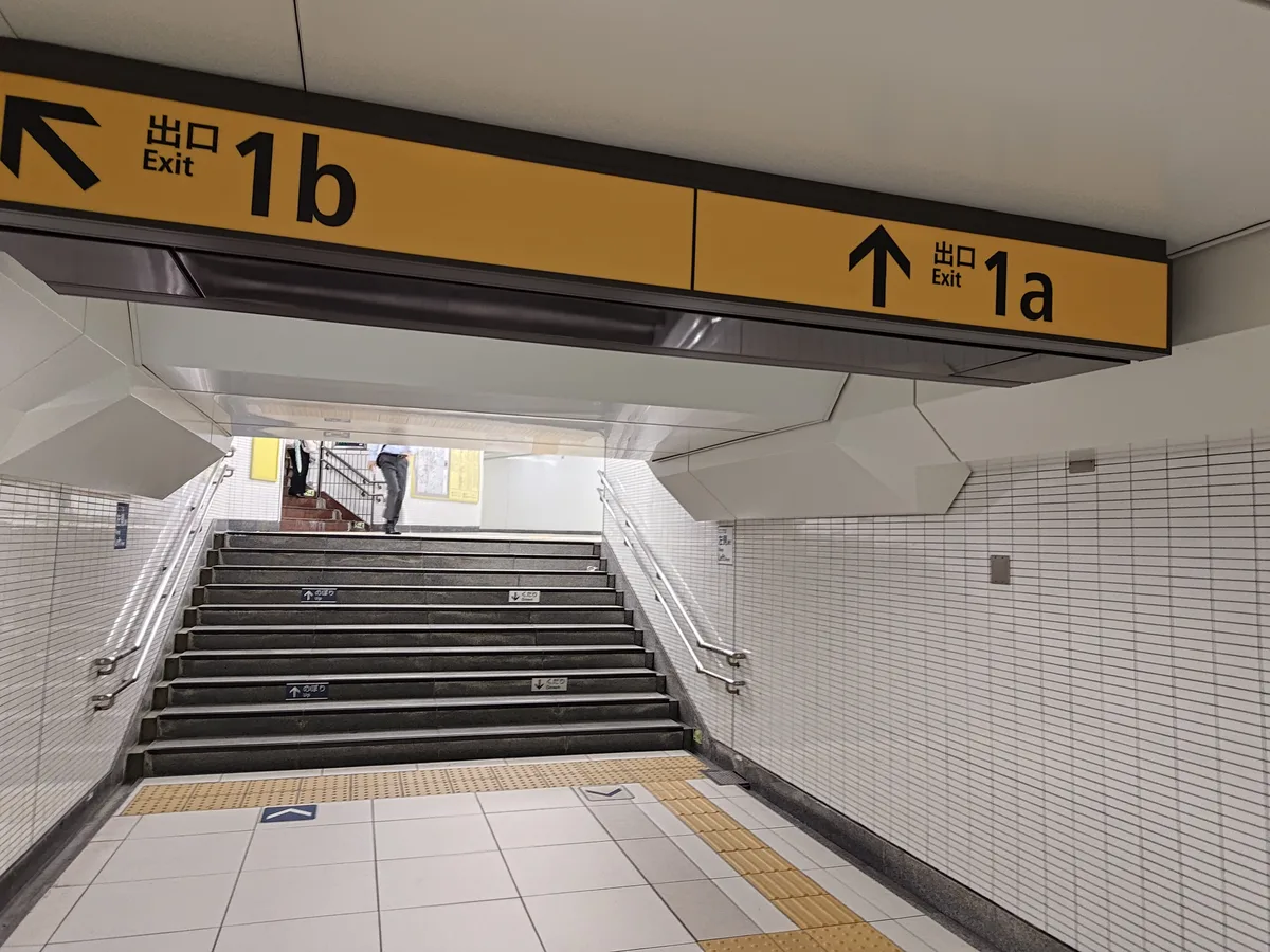 池袋站地下通道的出口指示牌。黃色招牌上並列顯示「出口 Exit 1a」（向上箭頭）和「出口 Exit 1b」（向左箭頭），前方可見通往地面的樓梯。 東京池袋 自動販賣機指南